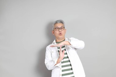 Doğu Asyalı bir adamın portresi gri saçlı ve gözlüklü, şık günlük kıyafetler giymiş, gri bir arka plana karşı duran.