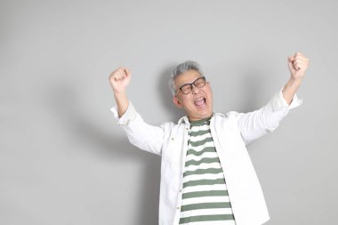 Doğu Asyalı bir adamın portresi gri saçlı ve gözlüklü, şık günlük kıyafetler giymiş, gri bir arka plana karşı duran.