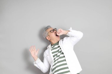 Doğu Asyalı bir adamın portresi gri saçlı ve gözlüklü, şık günlük kıyafetler giymiş, gri bir arka plana karşı duran.