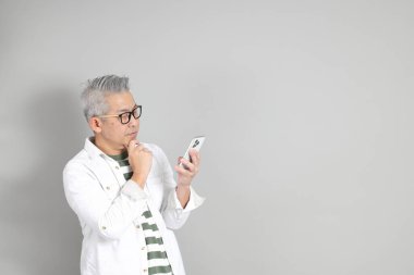 Doğu Asyalı bir adamın portresi gri saçlı ve gözlüklü, şık günlük kıyafetler giymiş, gri bir arka plana karşı duran.