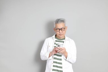 Doğu Asyalı bir adamın portresi gri saçlı ve gözlüklü, şık günlük kıyafetler giymiş, gri bir arka plana karşı duran.