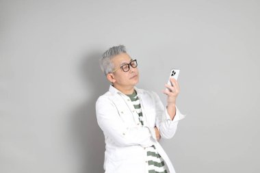 Doğu Asyalı bir adamın portresi gri saçlı ve gözlüklü, şık günlük kıyafetler giymiş, gri bir arka plana karşı duran.