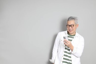 Doğu Asyalı bir adamın portresi gri saçlı ve gözlüklü, şık günlük kıyafetler giymiş, gri bir arka plana karşı duran.