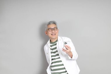 Doğu Asyalı bir adamın portresi gri saçlı ve gözlüklü, şık günlük kıyafetler giymiş, gri bir arka plana karşı duran.