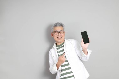 Doğu Asyalı bir adamın portresi gri saçlı ve gözlüklü, şık günlük kıyafetler giymiş, gri bir arka plana karşı duran.