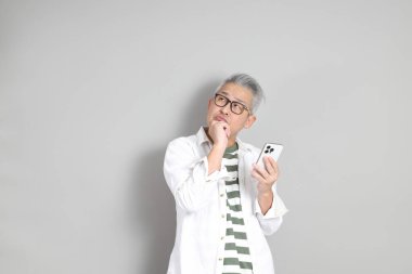 Doğu Asyalı bir adamın portresi gri saçlı ve gözlüklü, şık günlük kıyafetler giymiş, gri bir arka plana karşı duran.