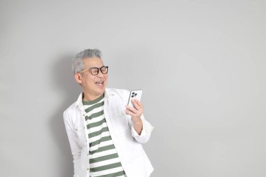 Doğu Asyalı bir adamın portresi gri saçlı ve gözlüklü, şık günlük kıyafetler giymiş, gri bir arka plana karşı duran.