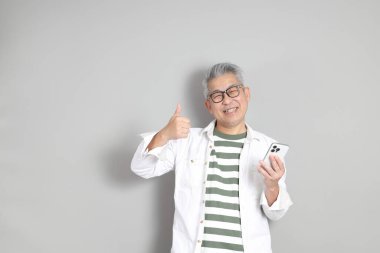 Doğu Asyalı bir adamın portresi gri saçlı ve gözlüklü, şık günlük kıyafetler giymiş, gri bir arka plana karşı duran.