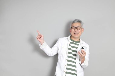 Doğu Asyalı bir adamın portresi gri saçlı ve gözlüklü, şık günlük kıyafetler giymiş, gri bir arka plana karşı duran.