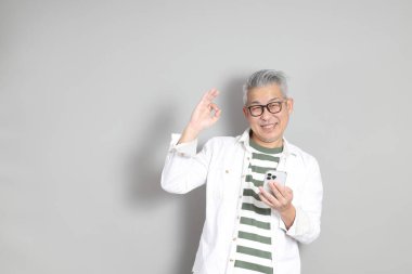 Doğu Asyalı bir adamın portresi gri saçlı ve gözlüklü, şık günlük kıyafetler giymiş, gri bir arka plana karşı duran.