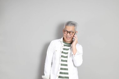 Doğu Asyalı bir adamın portresi gri saçlı ve gözlüklü, şık günlük kıyafetler giymiş, gri bir arka plana karşı duran.