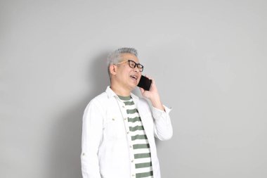 Doğu Asyalı bir adamın portresi gri saçlı ve gözlüklü, şık günlük kıyafetler giymiş, gri bir arka plana karşı duran.