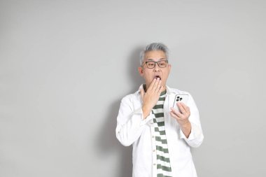 Doğu Asyalı bir adamın portresi gri saçlı ve gözlüklü, şık günlük kıyafetler giymiş, gri bir arka plana karşı duran.