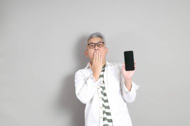 Doğu Asyalı bir adamın portresi gri saçlı ve gözlüklü, şık günlük kıyafetler giymiş, gri bir arka plana karşı duran.