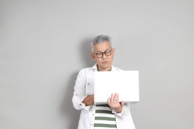 Doğu Asyalı bir adamın portresi gri saçlı ve gözlüklü, şık günlük kıyafetler giymiş, gri bir arka plana karşı duran.