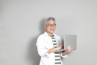 Doğu Asyalı bir adamın portresi gri saçlı ve gözlüklü, şık günlük kıyafetler giymiş, gri bir arka plana karşı duran.