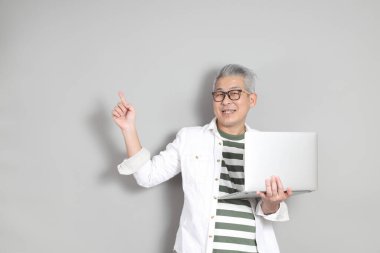 Doğu Asyalı bir adamın portresi gri saçlı ve gözlüklü, şık günlük kıyafetler giymiş, gri bir arka plana karşı duran.