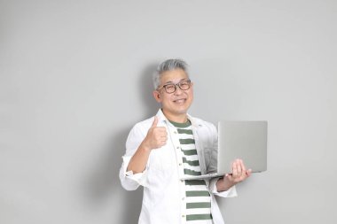 Doğu Asyalı bir adamın portresi gri saçlı ve gözlüklü, şık günlük kıyafetler giymiş, gri bir arka plana karşı duran.