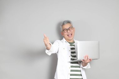 Doğu Asyalı bir adamın portresi gri saçlı ve gözlüklü, şık günlük kıyafetler giymiş, gri bir arka plana karşı duran.