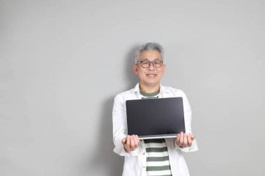 Doğu Asyalı bir adamın portresi gri saçlı ve gözlüklü, şık günlük kıyafetler giymiş, gri bir arka plana karşı duran.