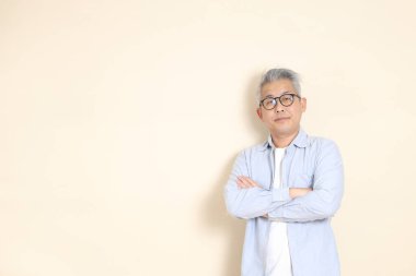 Doğu Asyalı bir adamın portresi gri saçlı ve gözlüklü, şık günlük kıyafetler giymiş, kahverengi arka planda duruyor..