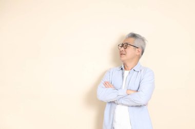 Doğu Asyalı bir adamın portresi gri saçlı ve gözlüklü, şık günlük kıyafetler giymiş, kahverengi arka planda duruyor..