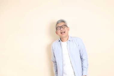 Doğu Asyalı bir adamın portresi gri saçlı ve gözlüklü, şık günlük kıyafetler giymiş, kahverengi arka planda duruyor..