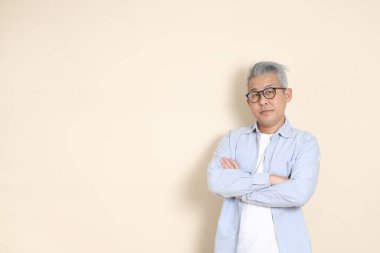 Doğu Asyalı bir adamın portresi gri saçlı ve gözlüklü, şık günlük kıyafetler giymiş, kahverengi arka planda duruyor..