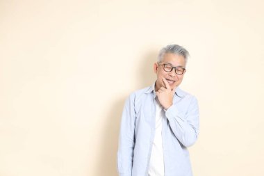 Doğu Asyalı bir adamın portresi gri saçlı ve gözlüklü, şık günlük kıyafetler giymiş, kahverengi arka planda duruyor..