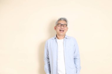 Doğu Asyalı bir adamın portresi gri saçlı ve gözlüklü, şık günlük kıyafetler giymiş, kahverengi arka planda duruyor..