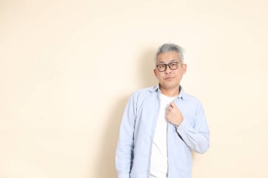 Doğu Asyalı bir adamın portresi gri saçlı ve gözlüklü, şık günlük kıyafetler giymiş, kahverengi arka planda duruyor..