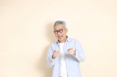 Doğu Asyalı bir adamın portresi gri saçlı ve gözlüklü, şık günlük kıyafetler giymiş, kahverengi arka planda duruyor..