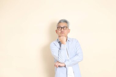 Doğu Asyalı bir adamın portresi gri saçlı ve gözlüklü, şık günlük kıyafetler giymiş, kahverengi arka planda duruyor..