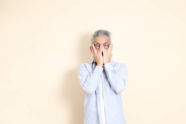 Doğu Asyalı bir adamın portresi gri saçlı ve gözlüklü, şık günlük kıyafetler giymiş, kahverengi arka planda duruyor..