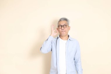 Doğu Asyalı bir adamın portresi gri saçlı ve gözlüklü, şık günlük kıyafetler giymiş, kahverengi arka planda duruyor..