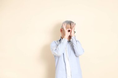 Doğu Asyalı bir adamın portresi gri saçlı ve gözlüklü, şık günlük kıyafetler giymiş, kahverengi arka planda duruyor..