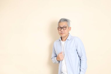 Doğu Asyalı bir adamın portresi gri saçlı ve gözlüklü, şık günlük kıyafetler giymiş, kahverengi arka planda duruyor..