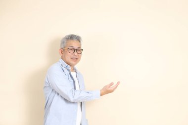 Doğu Asyalı bir adamın portresi gri saçlı ve gözlüklü, şık günlük kıyafetler giymiş, kahverengi arka planda duruyor..