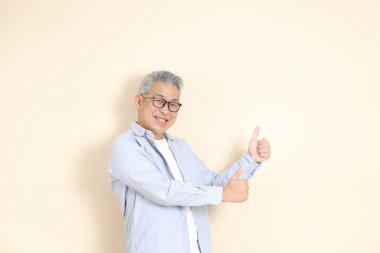 Doğu Asyalı bir adamın portresi gri saçlı ve gözlüklü, şık günlük kıyafetler giymiş, kahverengi arka planda duruyor..