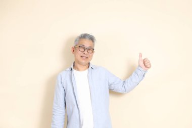 Doğu Asyalı bir adamın portresi gri saçlı ve gözlüklü, şık günlük kıyafetler giymiş, kahverengi arka planda duruyor..