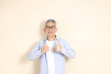 Doğu Asyalı bir adamın portresi gri saçlı ve gözlüklü, şık günlük kıyafetler giymiş, kahverengi arka planda duruyor..