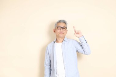 Doğu Asyalı bir adamın portresi gri saçlı ve gözlüklü, şık günlük kıyafetler giymiş, kahverengi arka planda duruyor..