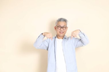 Doğu Asyalı bir adamın portresi gri saçlı ve gözlüklü, şık günlük kıyafetler giymiş, kahverengi arka planda duruyor..