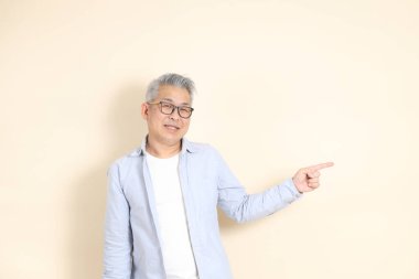 Doğu Asyalı bir adamın portresi gri saçlı ve gözlüklü, şık günlük kıyafetler giymiş, kahverengi arka planda duruyor..