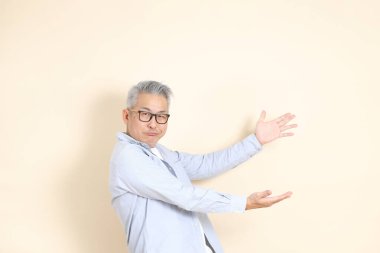 Doğu Asyalı bir adamın portresi gri saçlı ve gözlüklü, şık günlük kıyafetler giymiş, kahverengi arka planda duruyor..