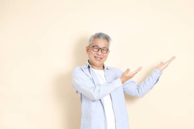 Doğu Asyalı bir adamın portresi gri saçlı ve gözlüklü, şık günlük kıyafetler giymiş, kahverengi arka planda duruyor..