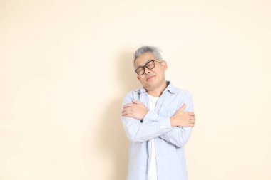 Doğu Asyalı bir adamın portresi gri saçlı ve gözlüklü, şık günlük kıyafetler giymiş, kahverengi arka planda duruyor..