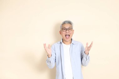 Doğu Asyalı bir adamın portresi gri saçlı ve gözlüklü, şık günlük kıyafetler giymiş, kahverengi arka planda duruyor..