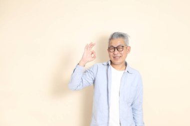 Doğu Asyalı bir adamın portresi gri saçlı ve gözlüklü, şık günlük kıyafetler giymiş, kahverengi arka planda duruyor..