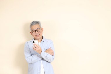 Doğu Asyalı bir adamın portresi gri saçlı ve gözlüklü, şık günlük kıyafetler giymiş, kahverengi arka planda duruyor..